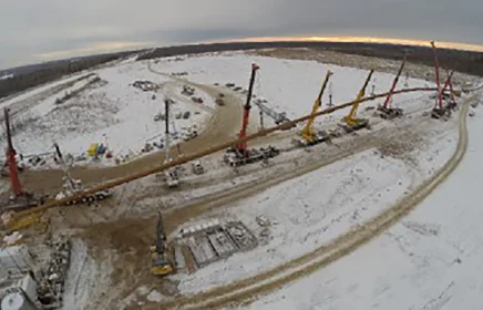 Large cranes hold a long, large diameter pipe in a snowy environment.