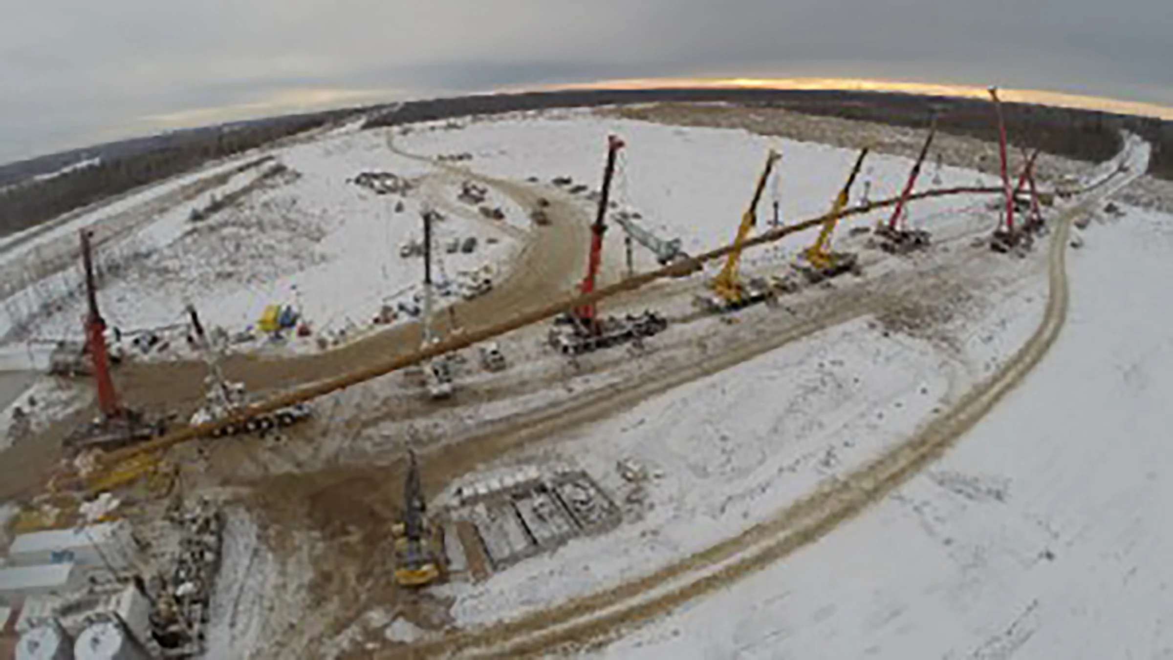 Large cranes hold a long, large diameter pipe in a snowy environment.