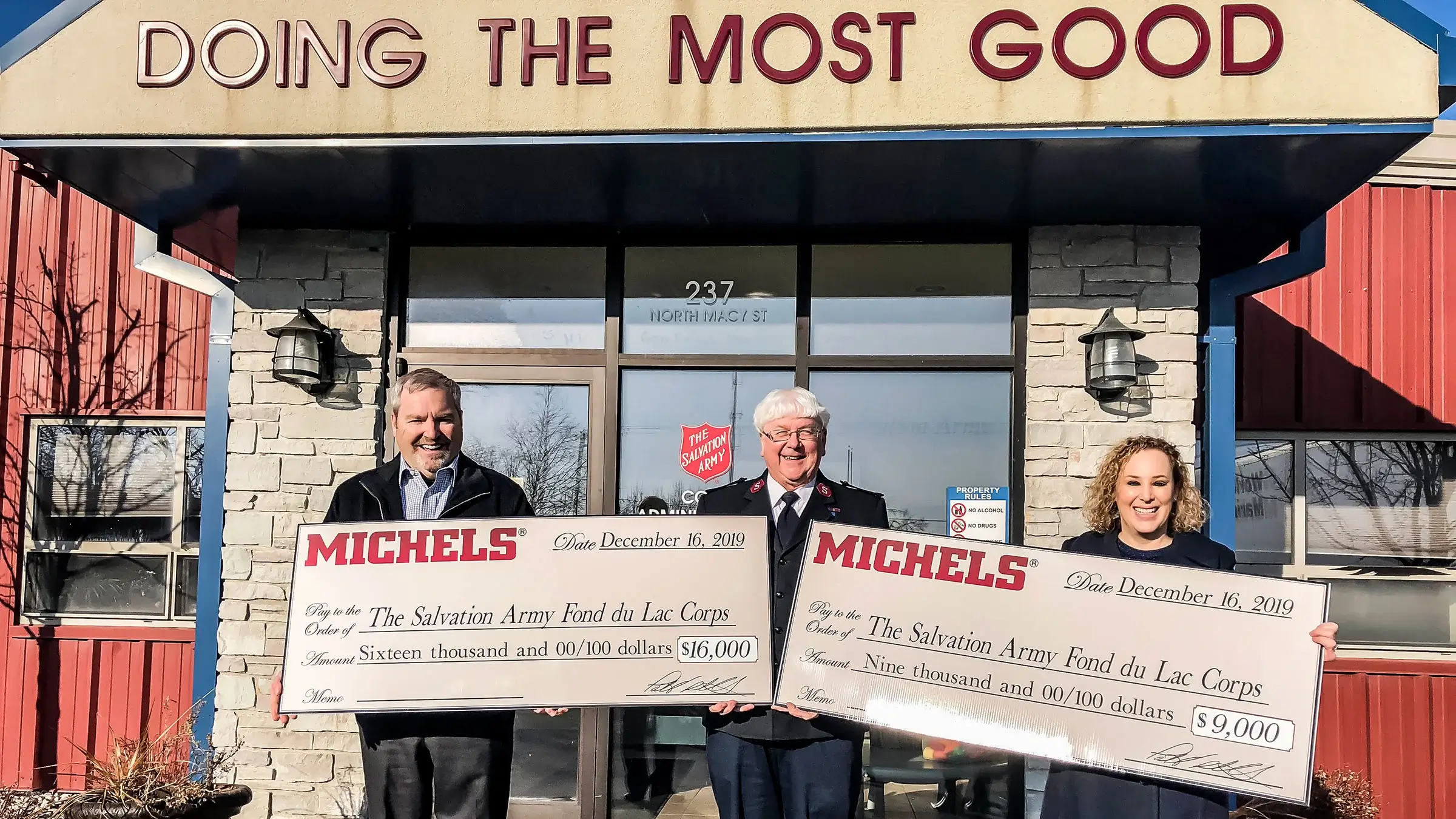 Michels stands outside of Salvation Army building with large checks