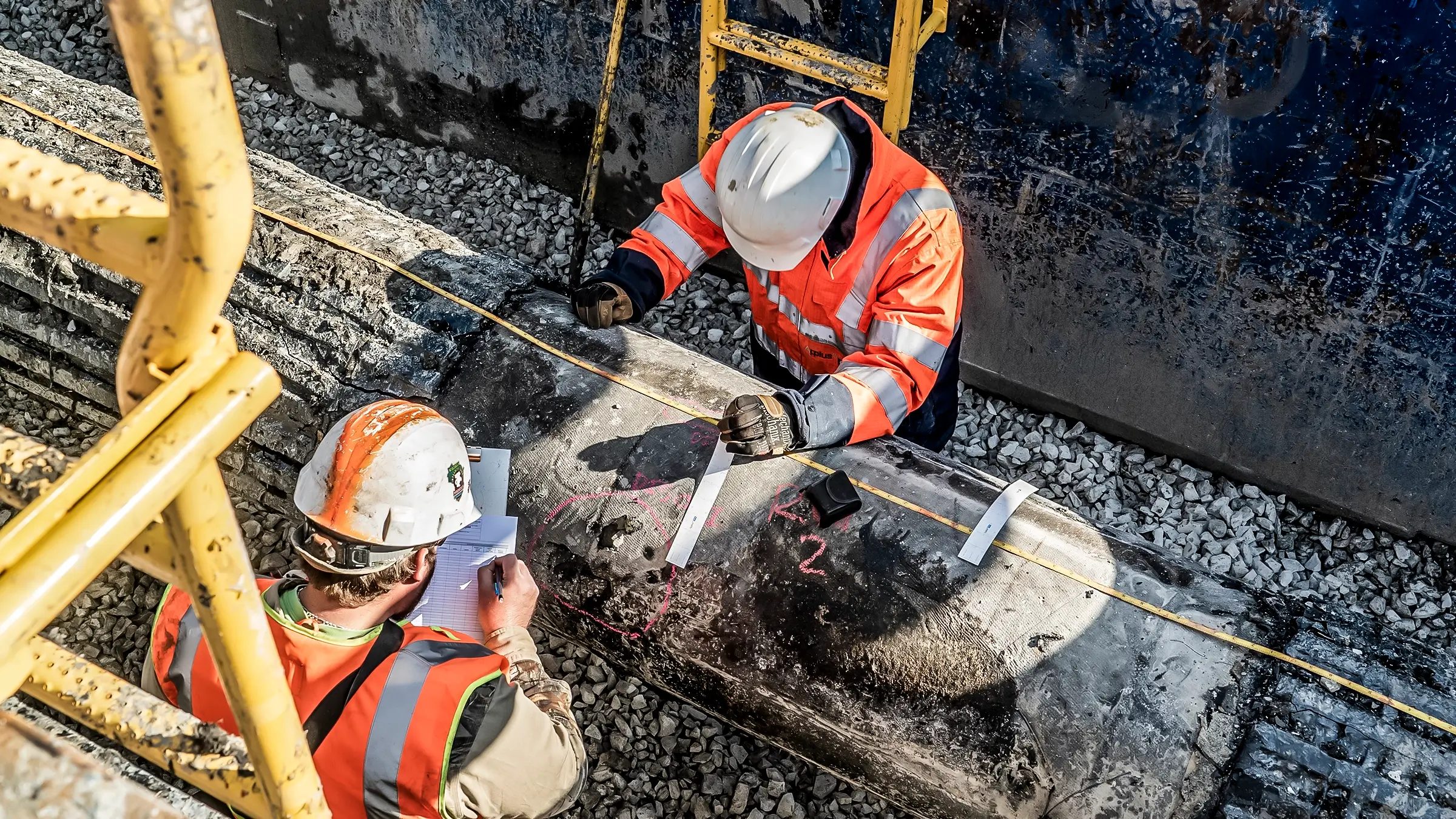 Two crew members take measurements of a pipeline in a trench.