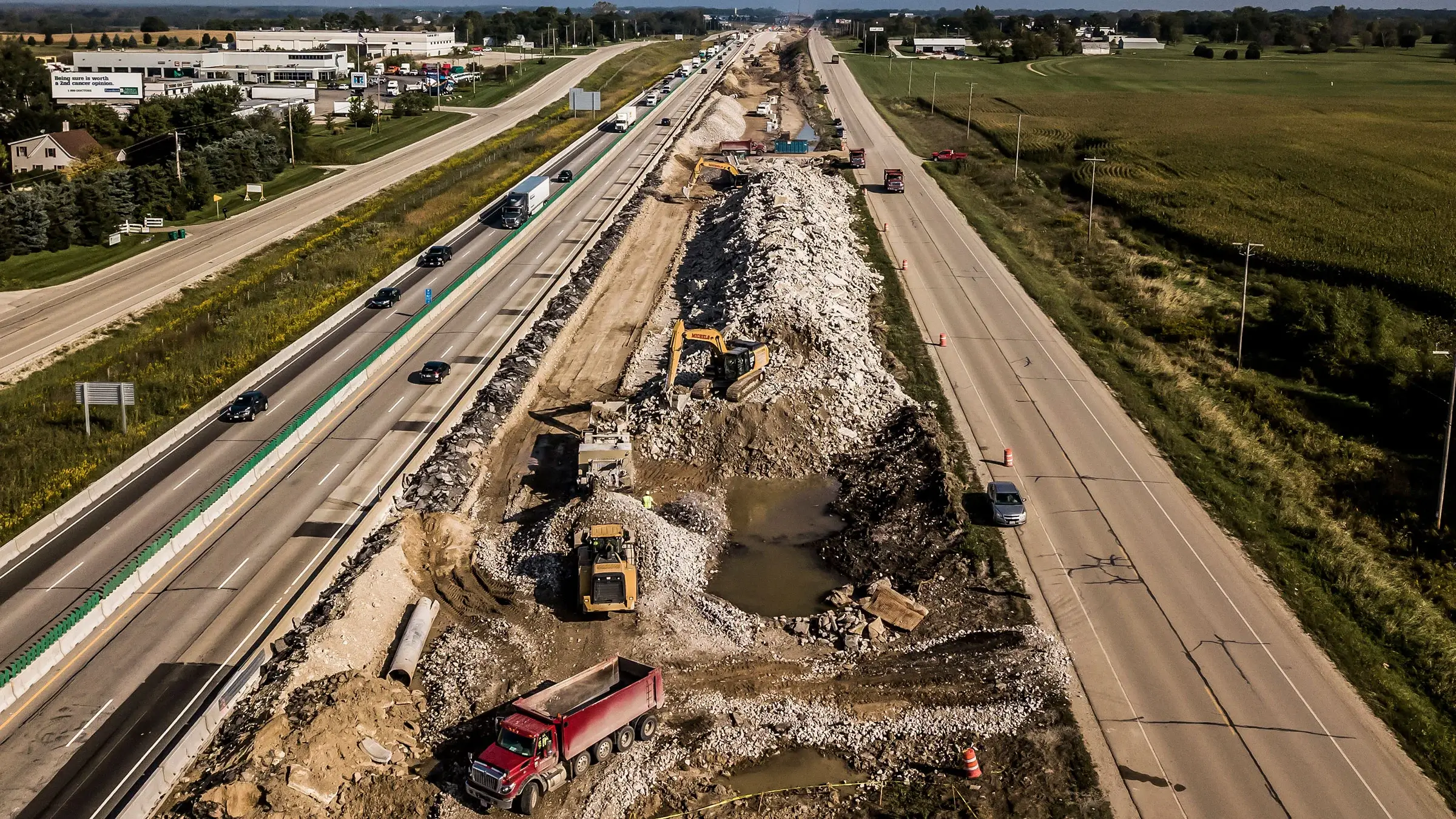 Excavators and dump trucks work to remove crushed aggregates from between two busy highways