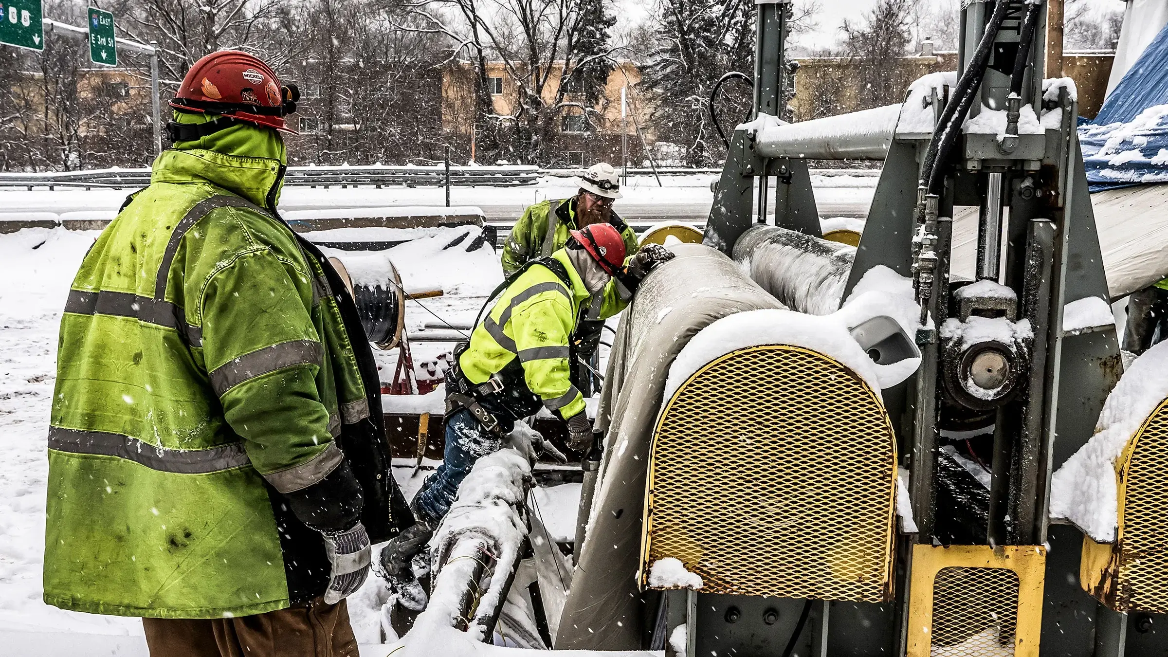 Three crew members operate on a sewer rehabiltion job in the snow near a busy freeway