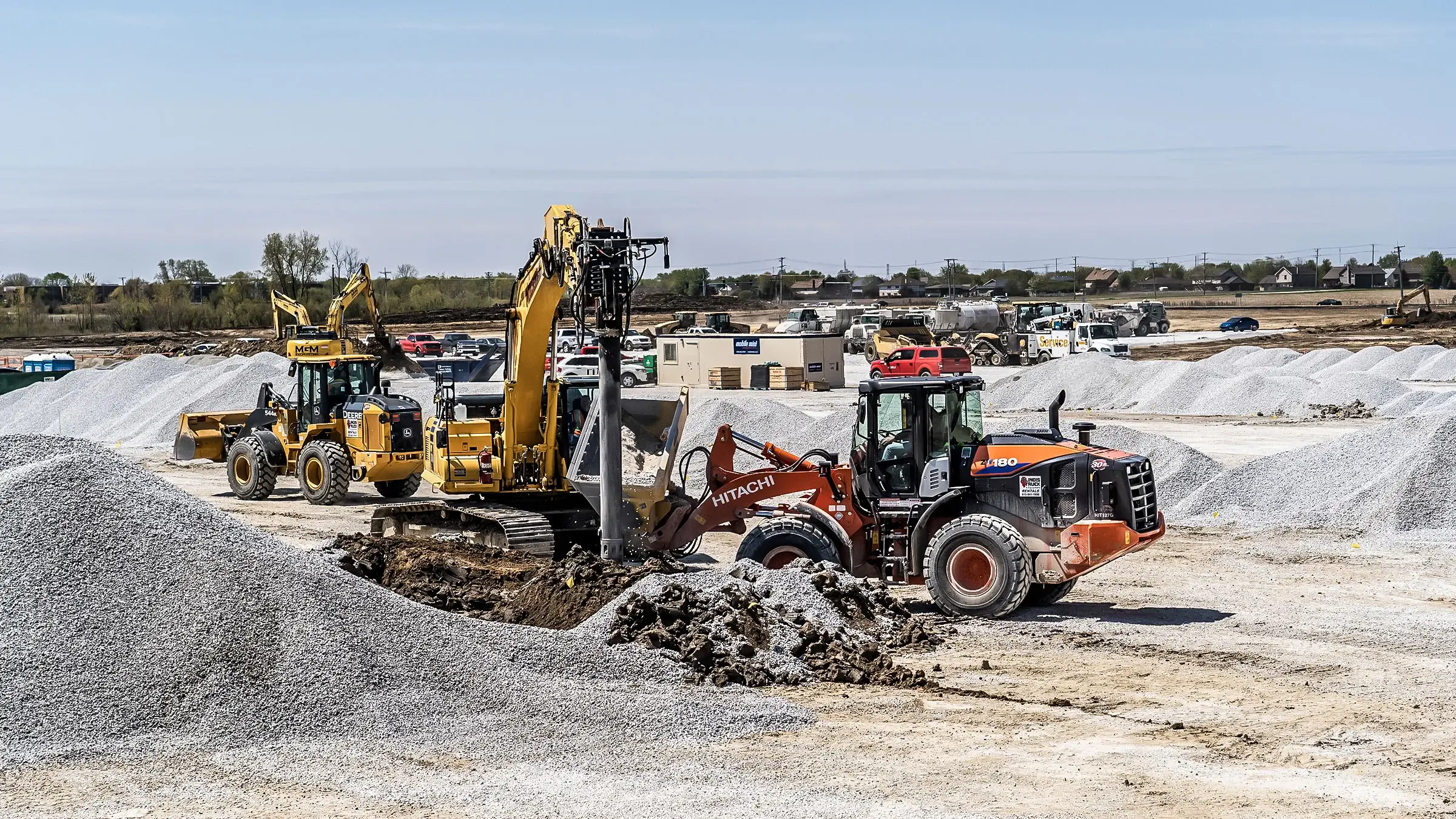 An excavator and backhoe assist in the installation of a ground improvement foundations job with many piles of aggregate nearby