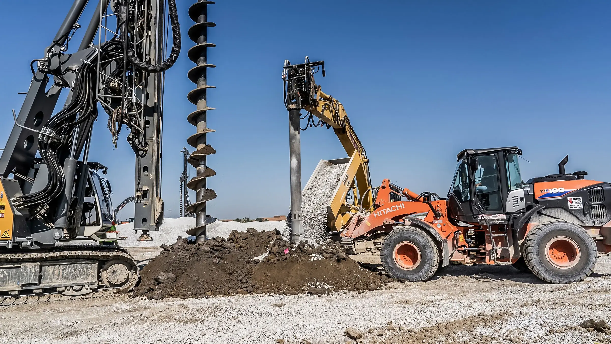 A drill rig, backhoe, and excavator all work at the same spot on a foundation