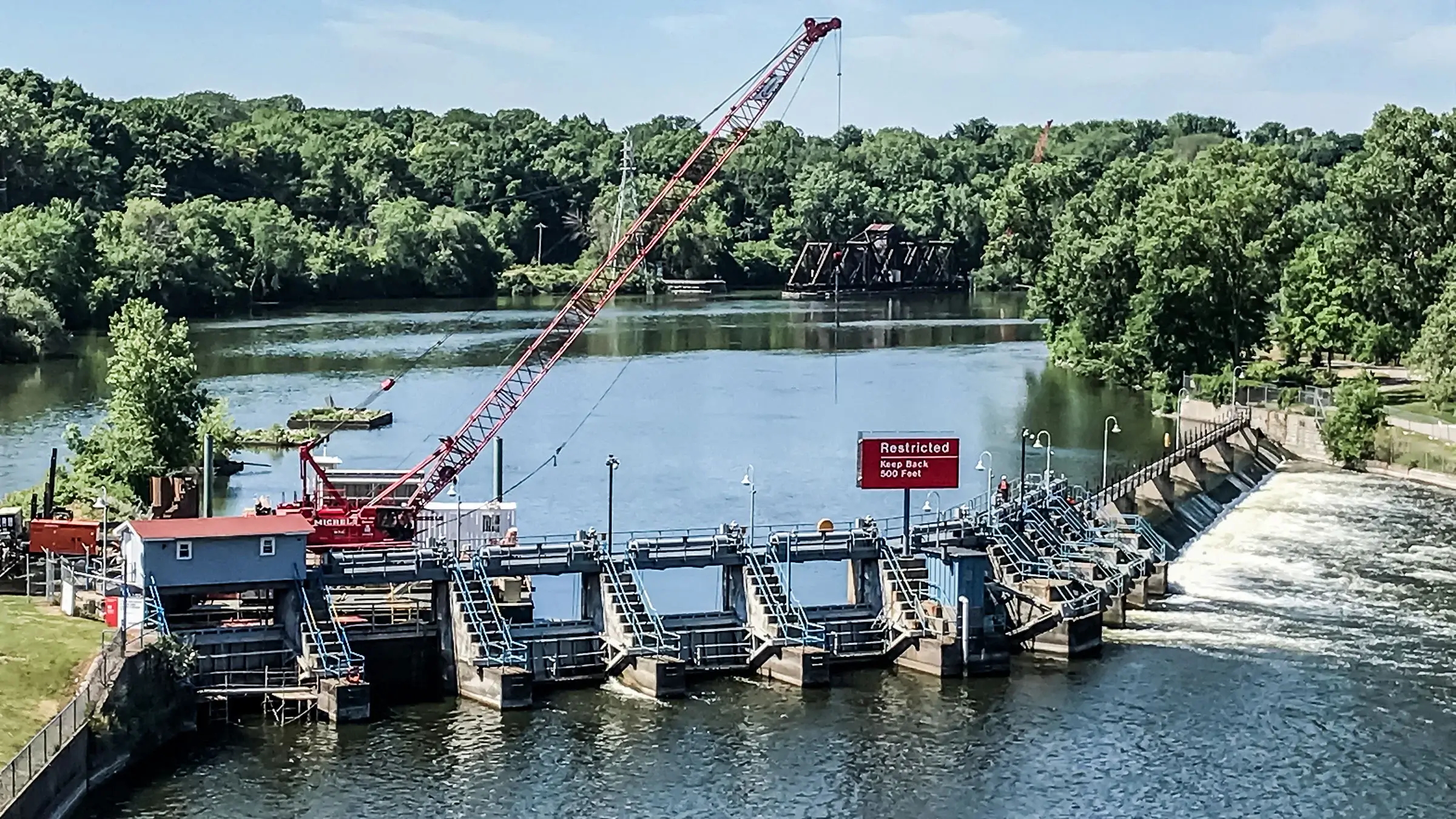 Crane operating over a dam on a sunny day with trees in the background.