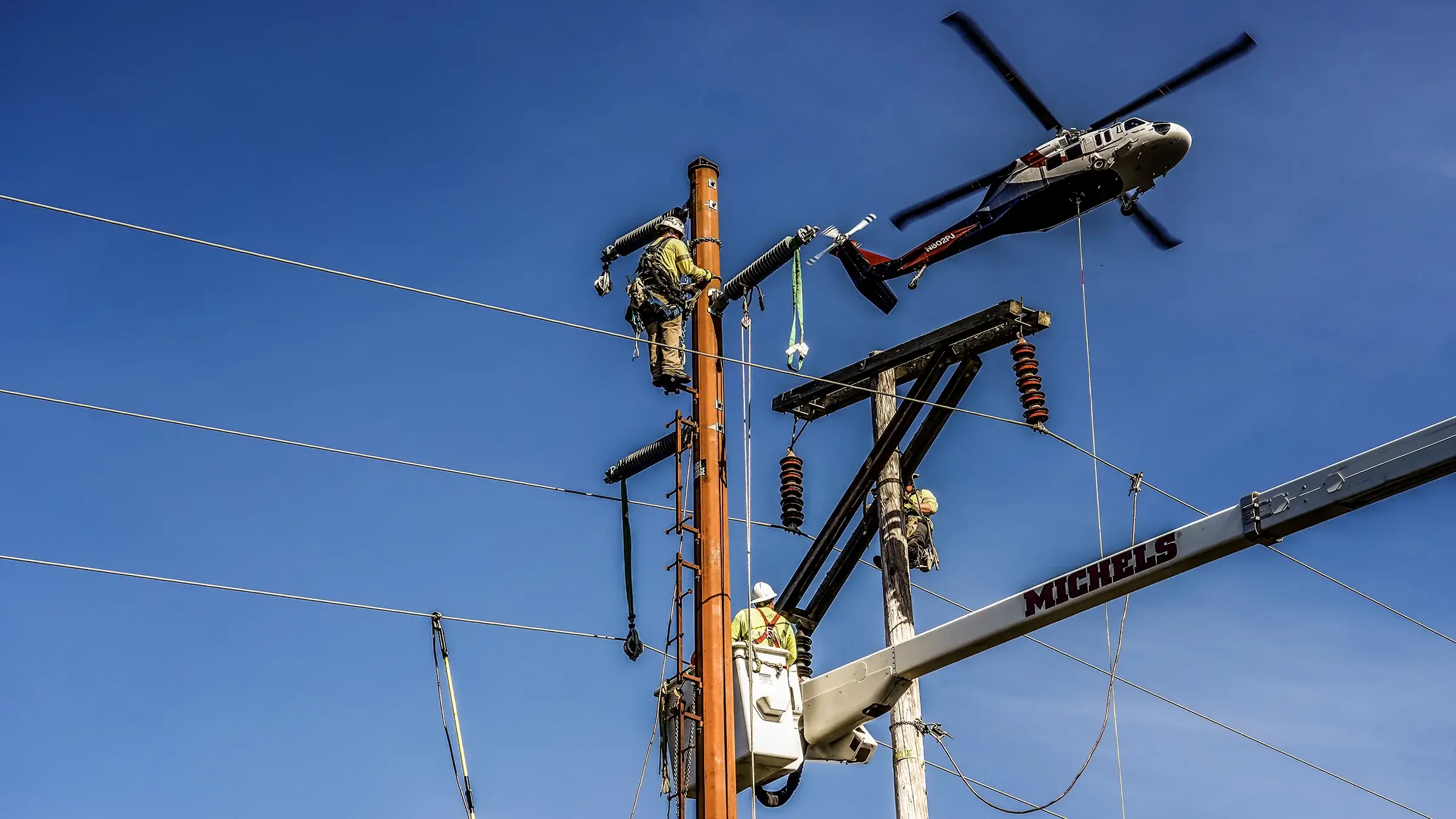 Electrical line workers work on a pole and from a bucket truck as a helicopter flies overhead