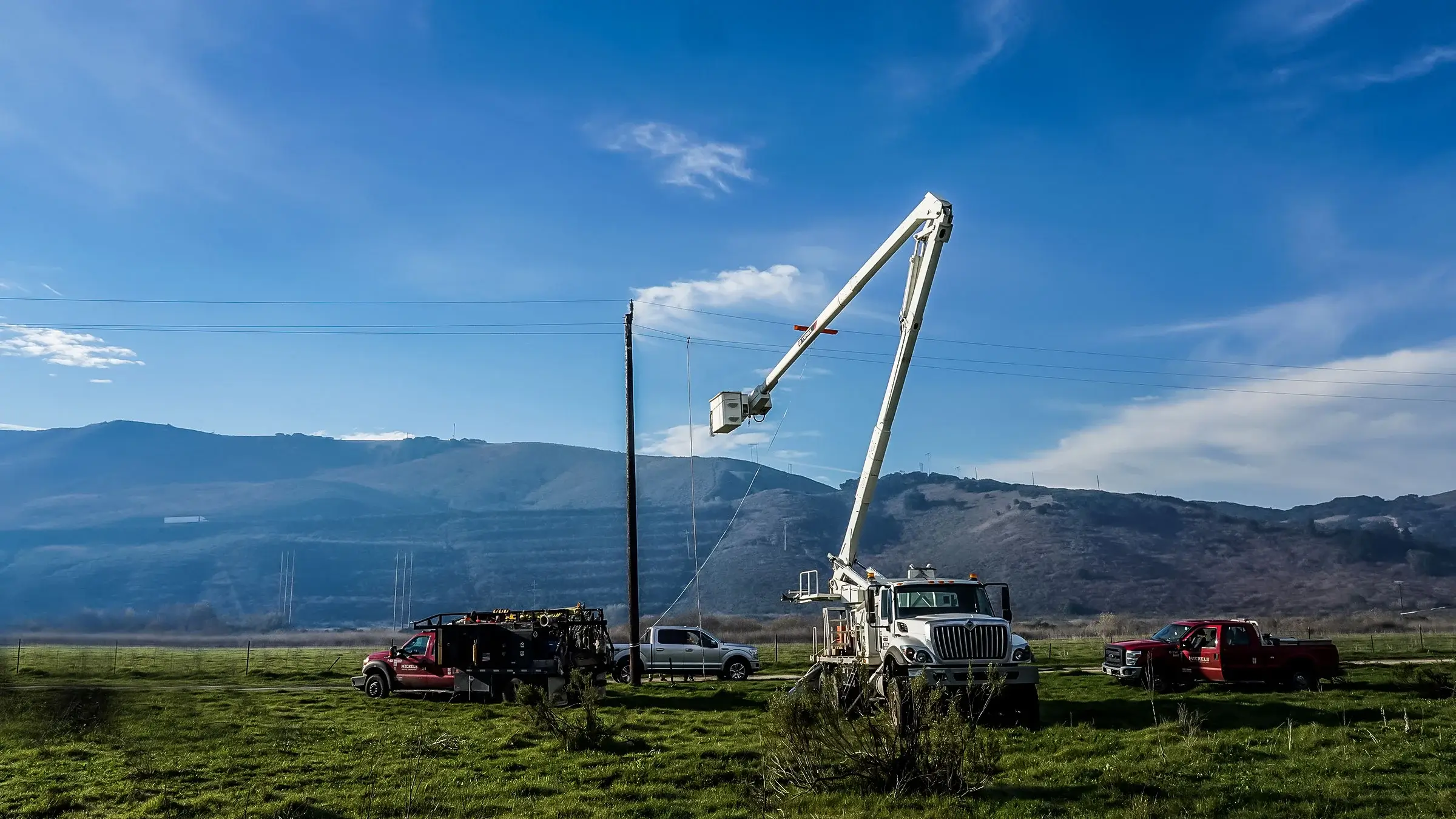 Power crew uses a bucket truck to work on power lines next to a grassy hillside.