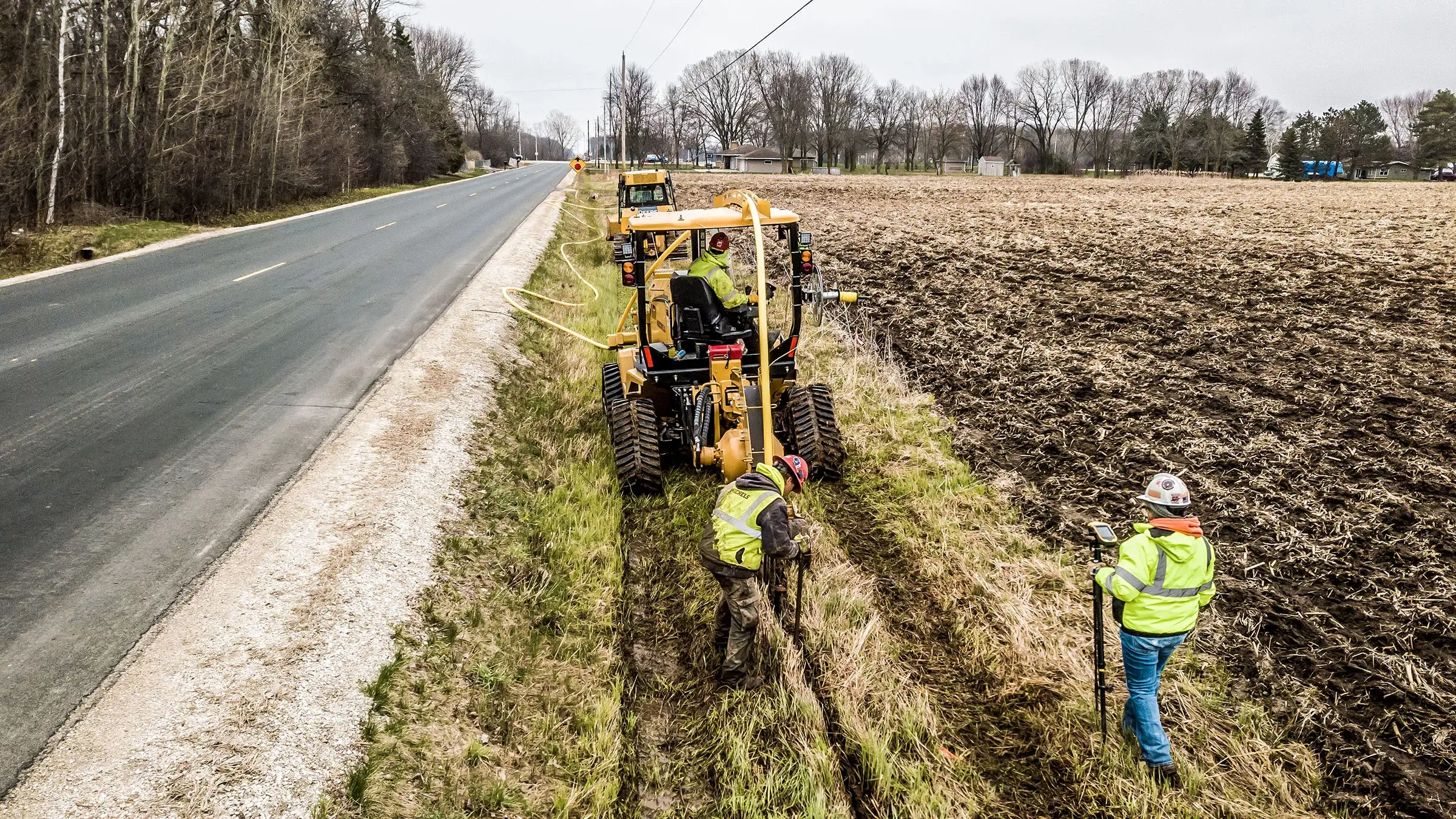 A utlity services crew operates a machine as it lays pipeline in ground near roadside.