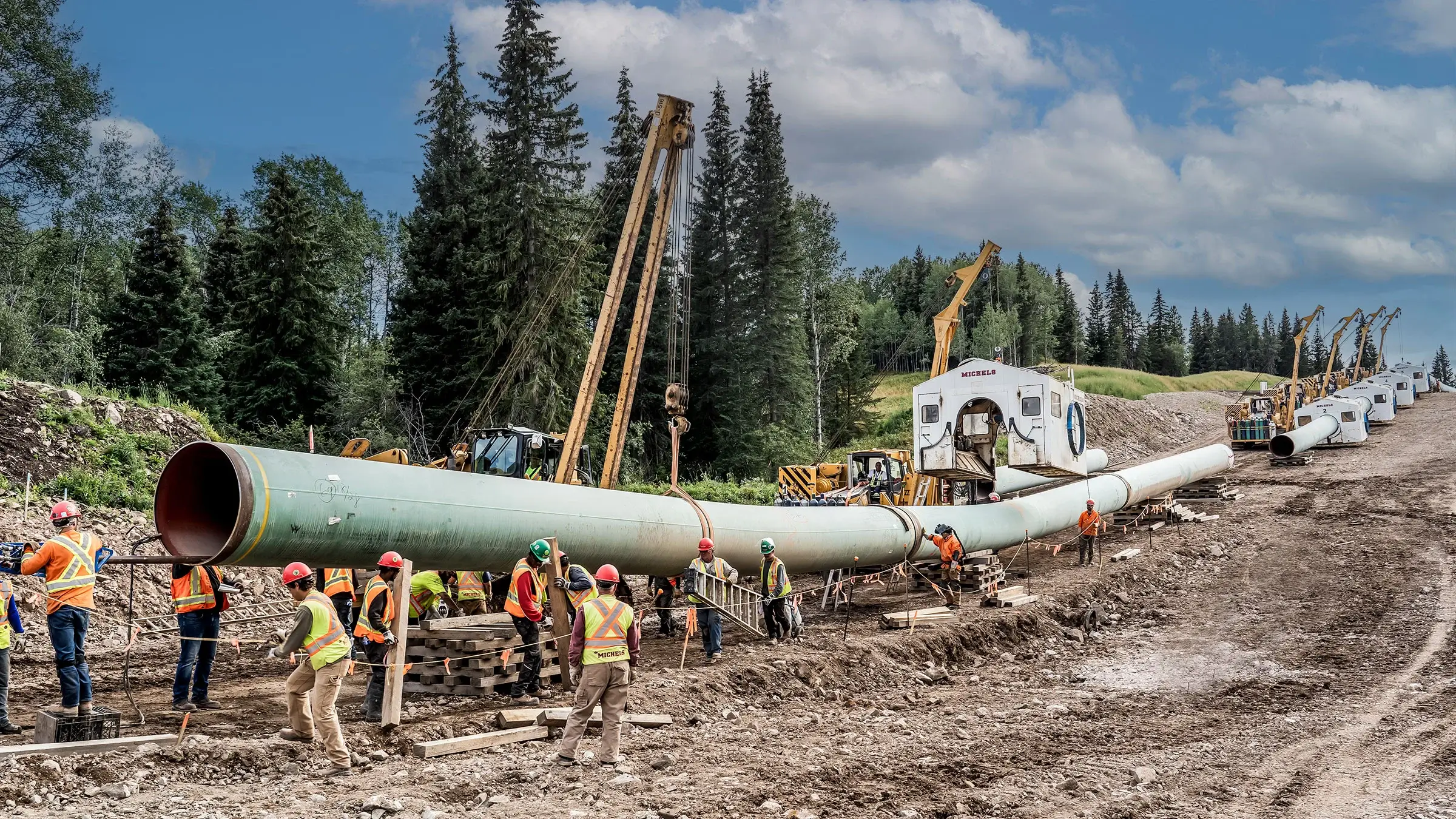 Workers operate along a long suspended pipeline in the forest.