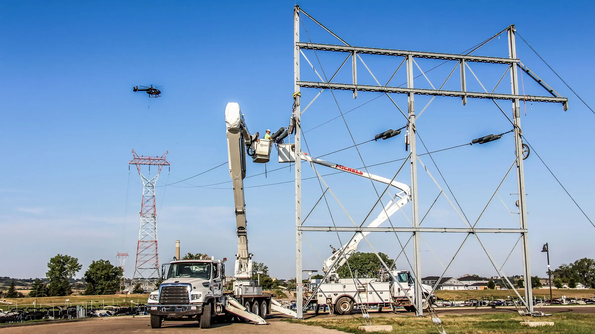 Two Michels Power bucket trucks operate with a helicopter nearby on large electrical structures.