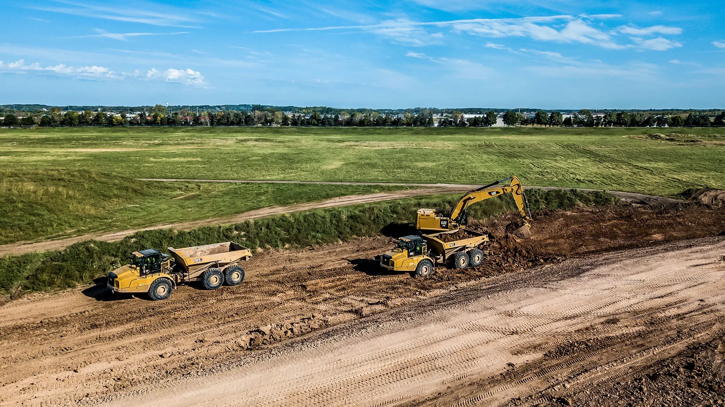 An excavator and several dumptrucks operate near a dirt road side, surrounded by green fields.