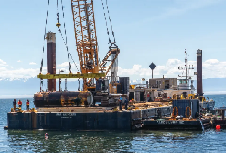 A large crane holding a piece of tunneling equipment on a water barge.