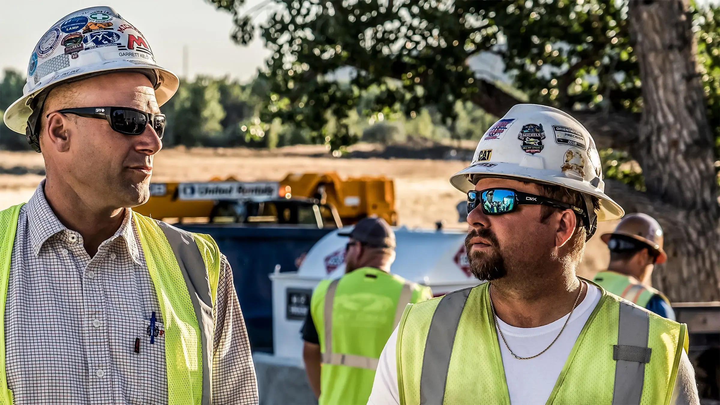 Two pipeline workers wearing hard hats and safety glasses talk to one another