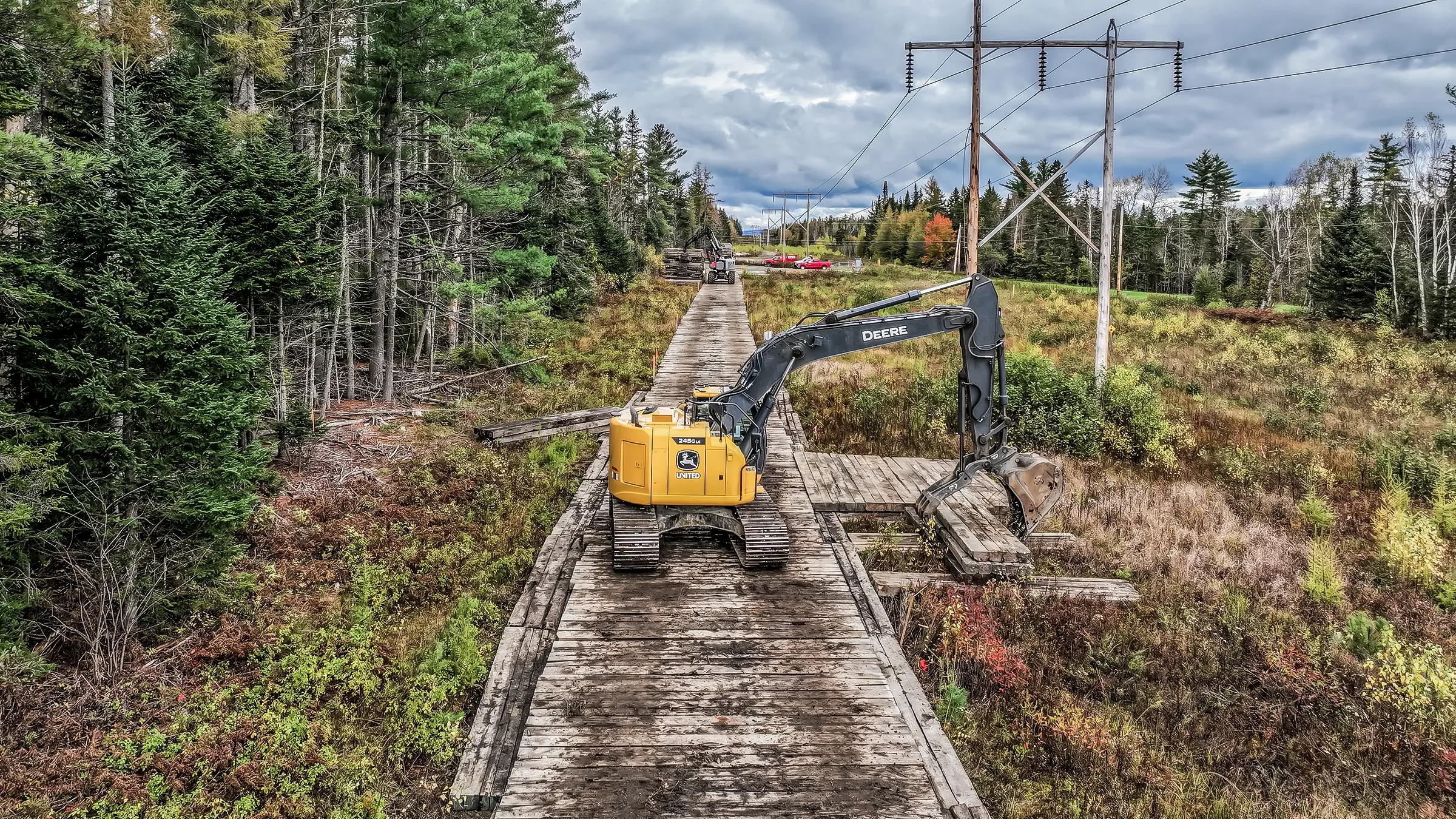 An excavator works along construction matting in a forest area.