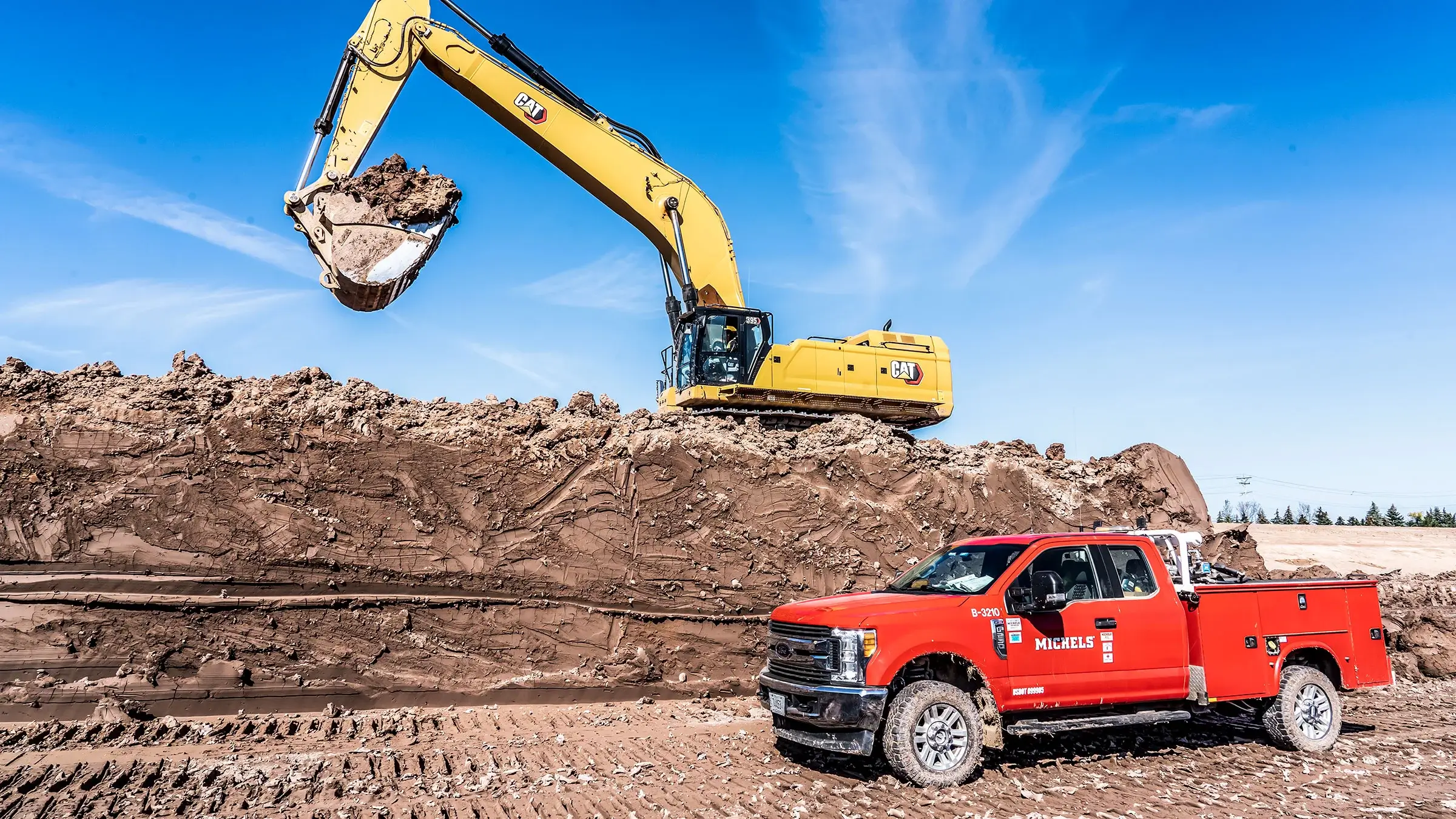 An excavator sits upon a pile of dirt near a red Michels pickup truck.