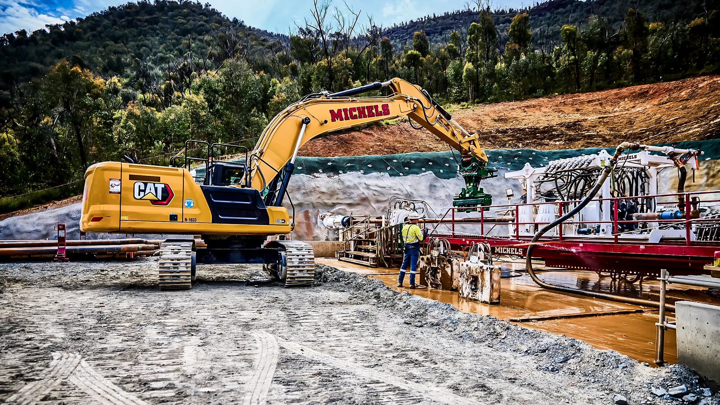 An HDD rig and excavator work side by side in a rural forested region of Australia.