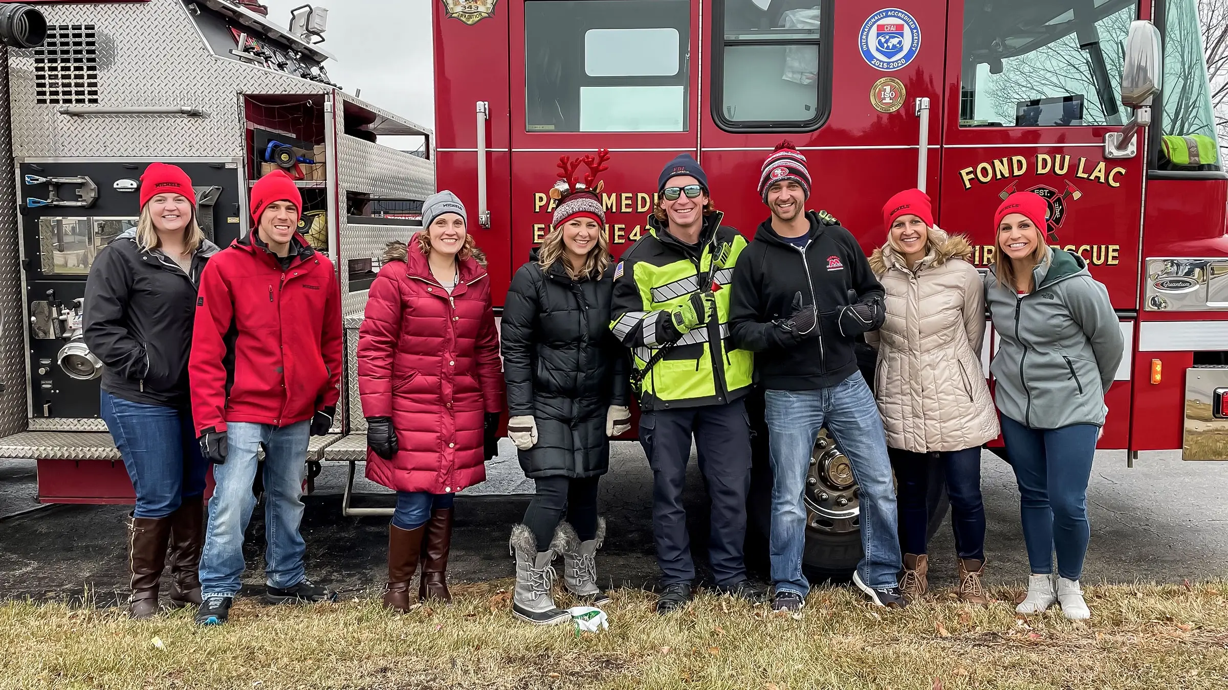 Group of employees and firefighters stand in front of a fire truck while ringing the bell for the salvation army.