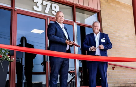 Two men stand outside a grand opening of an office with ribbon and scissors