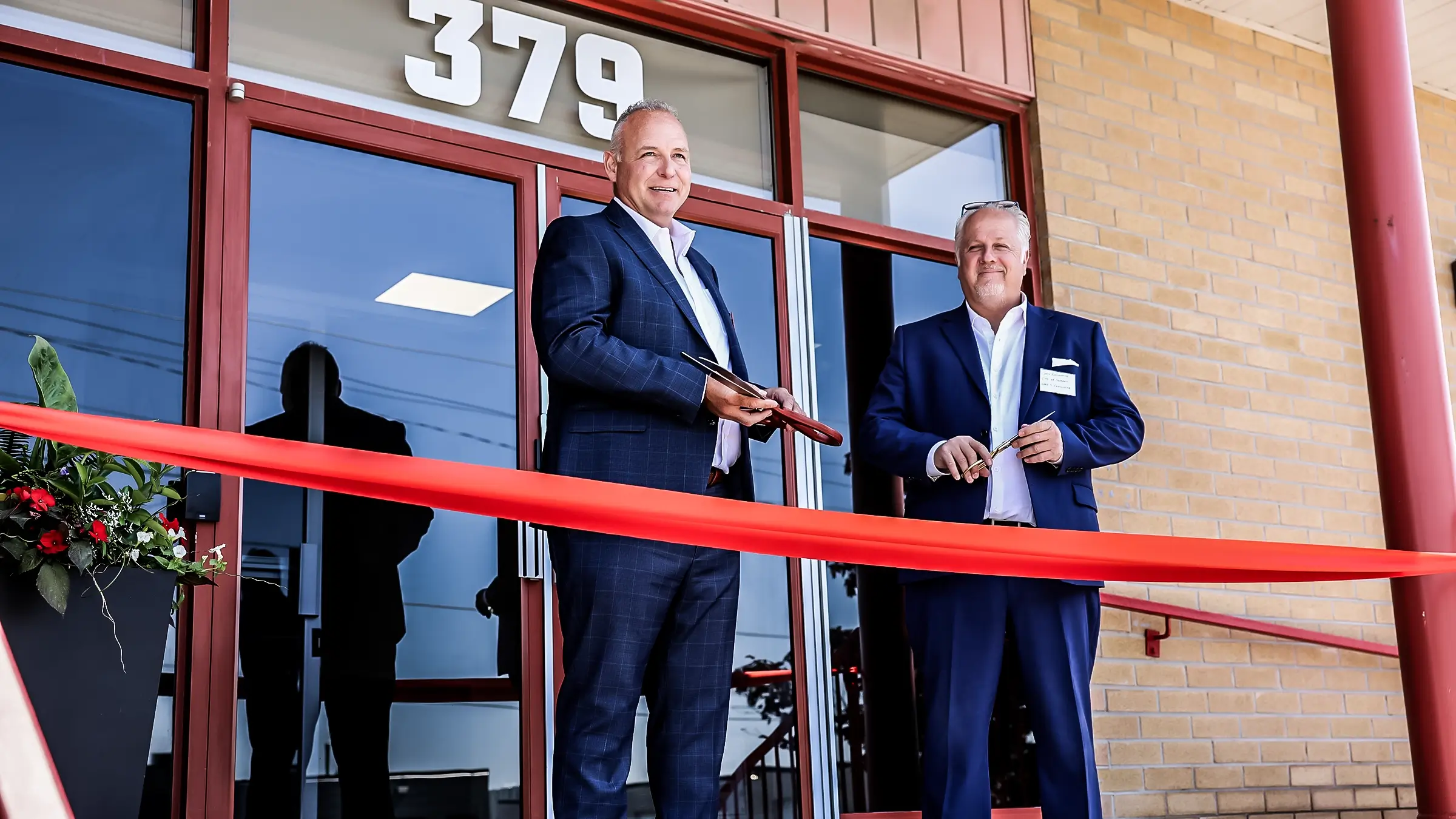 Two men stand outside a grand opening of an office with ribbon and scissors