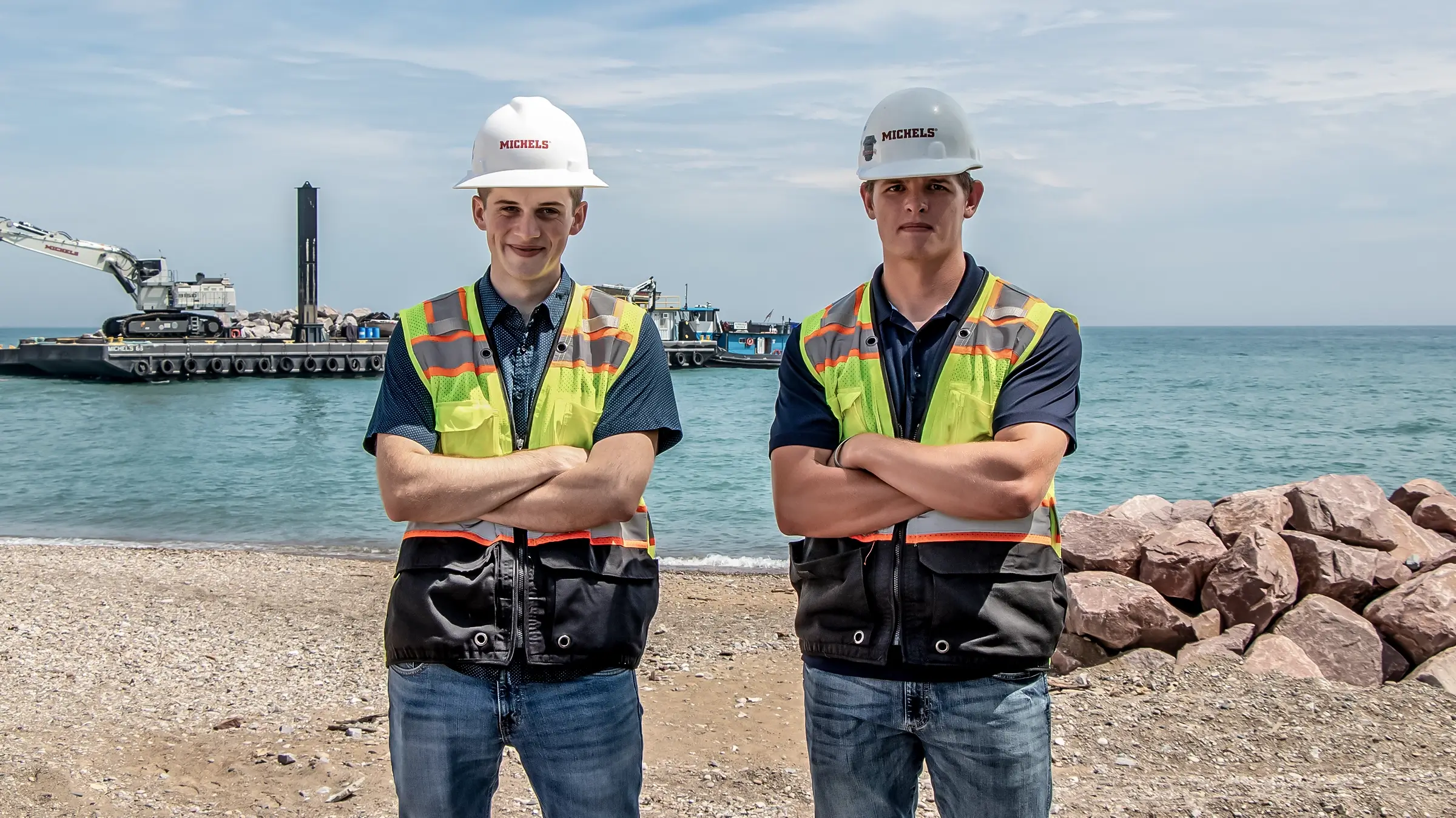 Two interns wearing PPE stand near a marine jobsite.