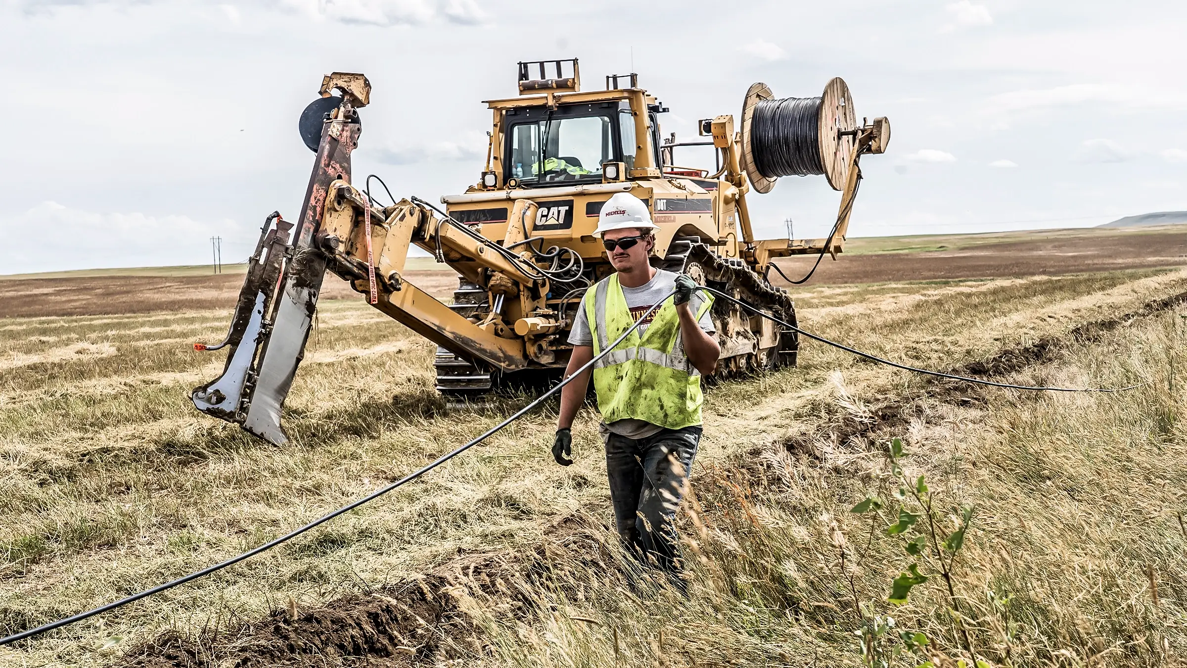 A crew member carries a coil of cable through a field near the plowing machine.
