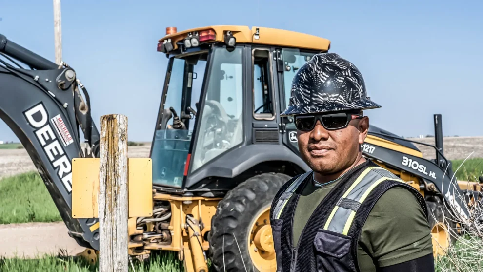 Man in rural field stands next to utility excavator.