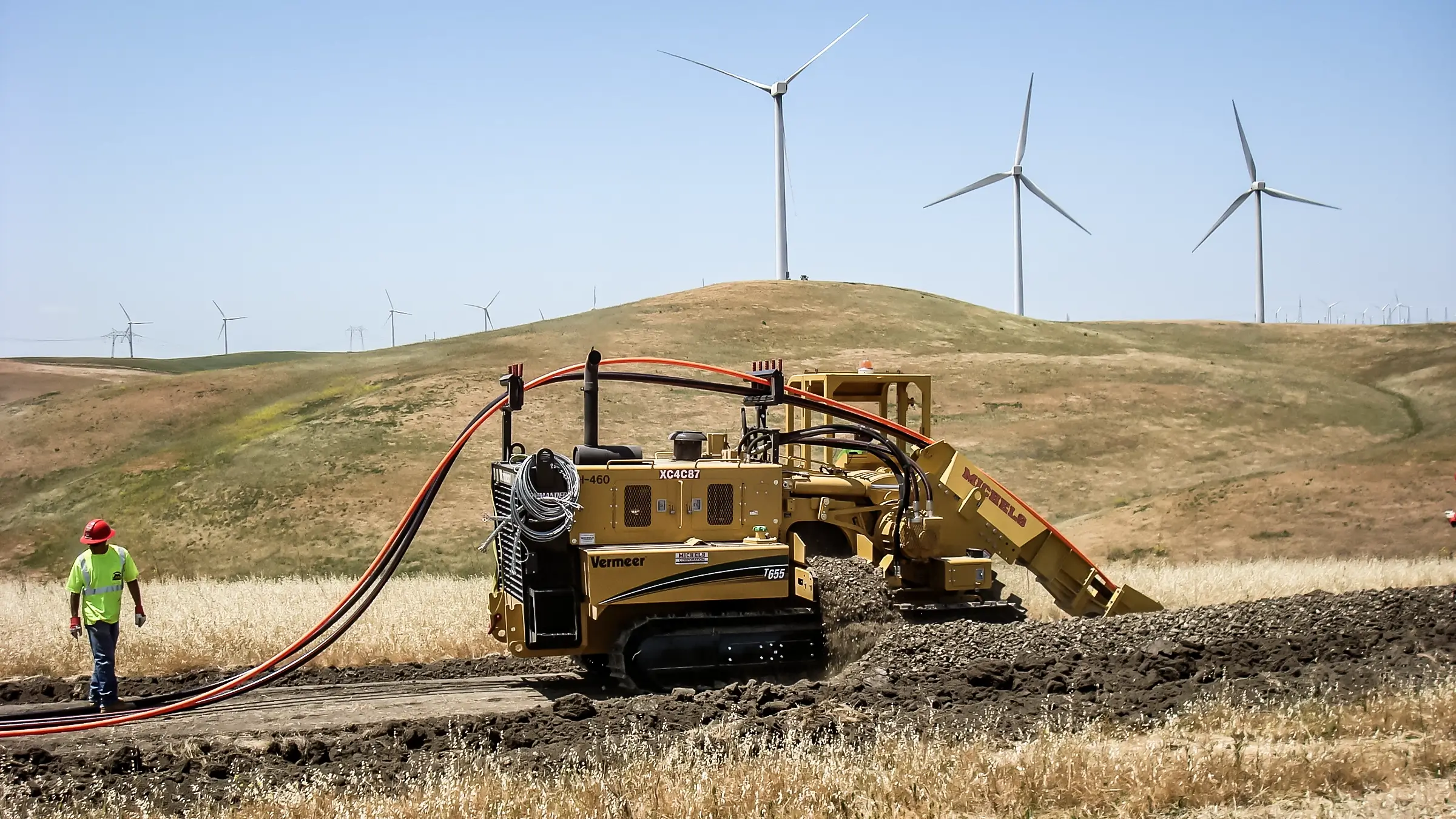 A crew member and cable installing machinery install cable near wind mills.